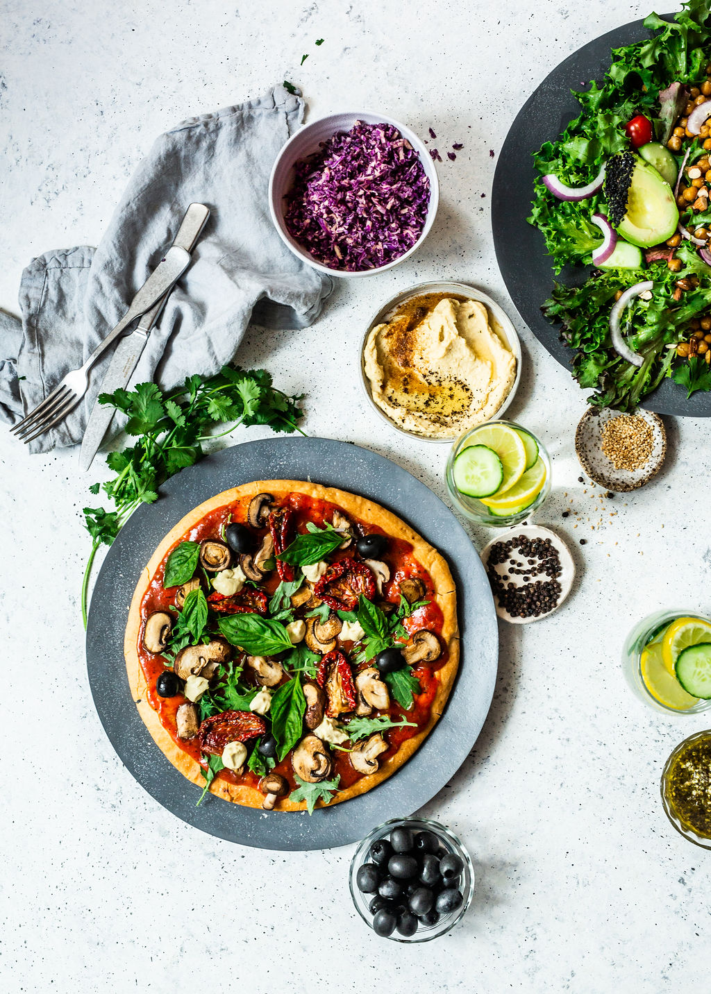 An array of food in a flatlay image. A pizza surrounded by hummus, coriander, lemons, cucumber, olives and a kale salad featuring some avocado and red onion.